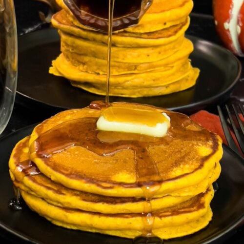 Stack of fluffy pumpkin pancakes with butter and maple syrup, golden edges on a dark plate.