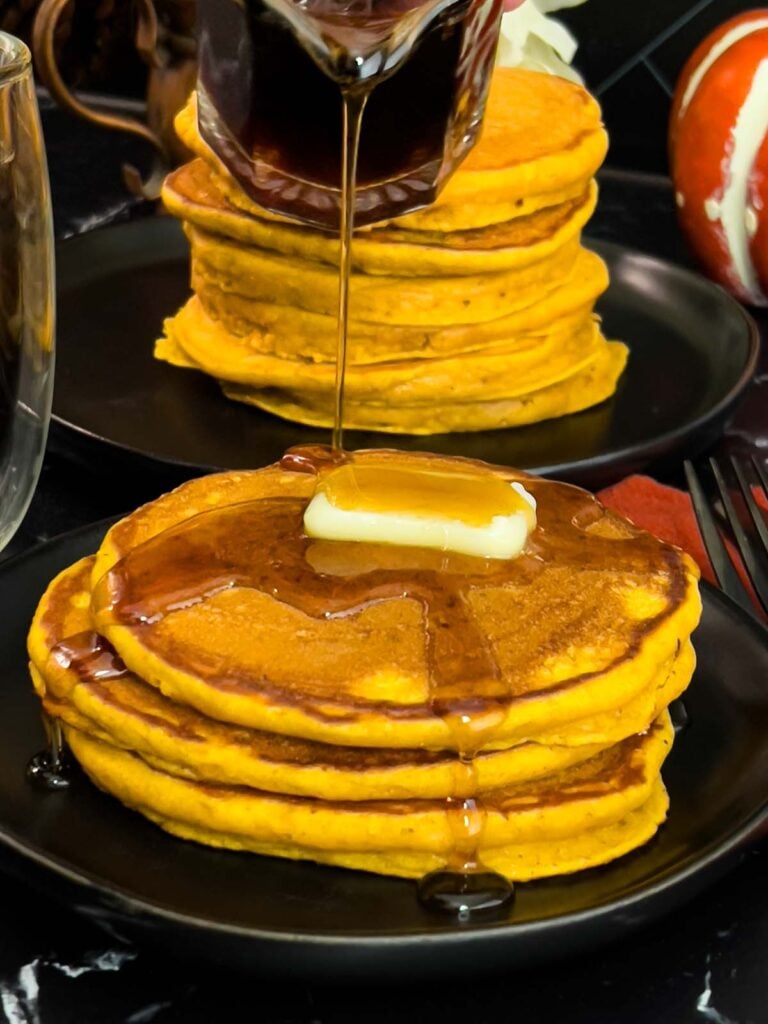 Stack of fluffy pumpkin pancakes with butter and maple syrup, golden edges on a dark plate.