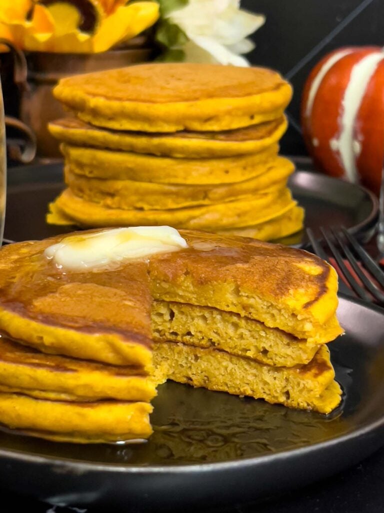Stack of fluffy pumpkin pancakes with butter and maple syrup, golden edges on a dark plate.