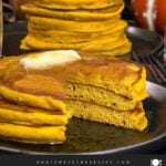 Stack of fluffy pumpkin pancakes with butter and maple syrup, golden edges on a dark plate.