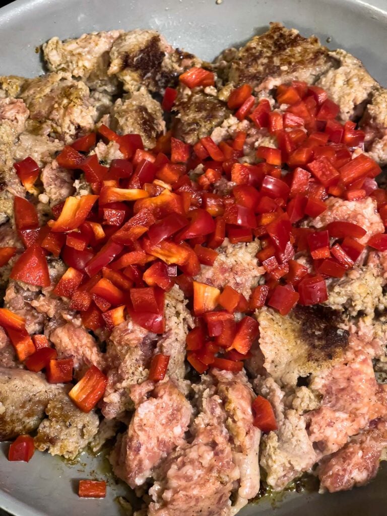 Diced red bell peppers added to browned sausage in skillet.