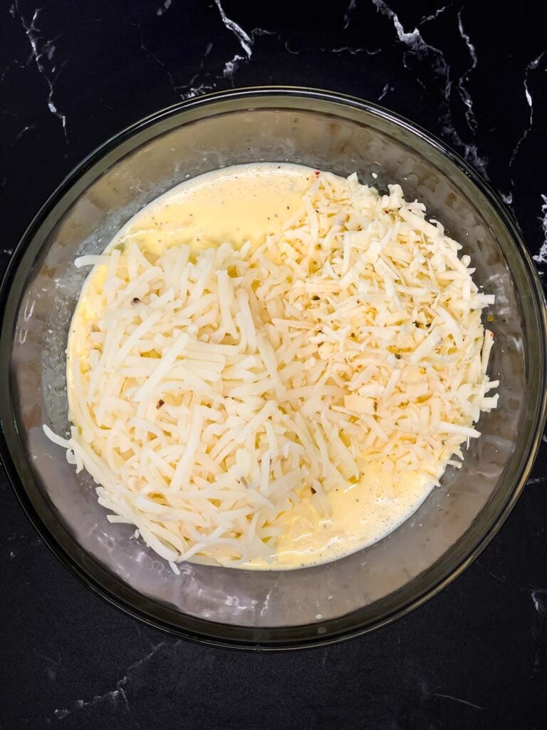 Whisked egg mixture with shredded hash browns and cheese added on top in a mixing bowl.