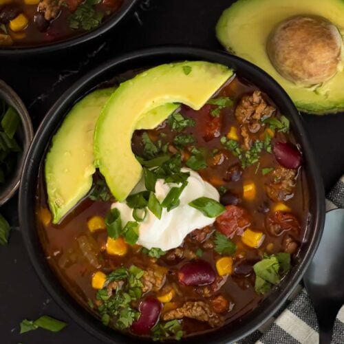 Bowls of taco soup in dark bowls with many garnishes.