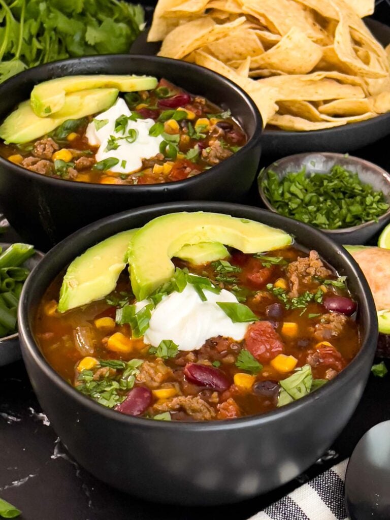 Bowls of taco soup in dark bowls with many garnishes.