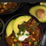 Bowls of taco soup in dark bowls with many garnishes.