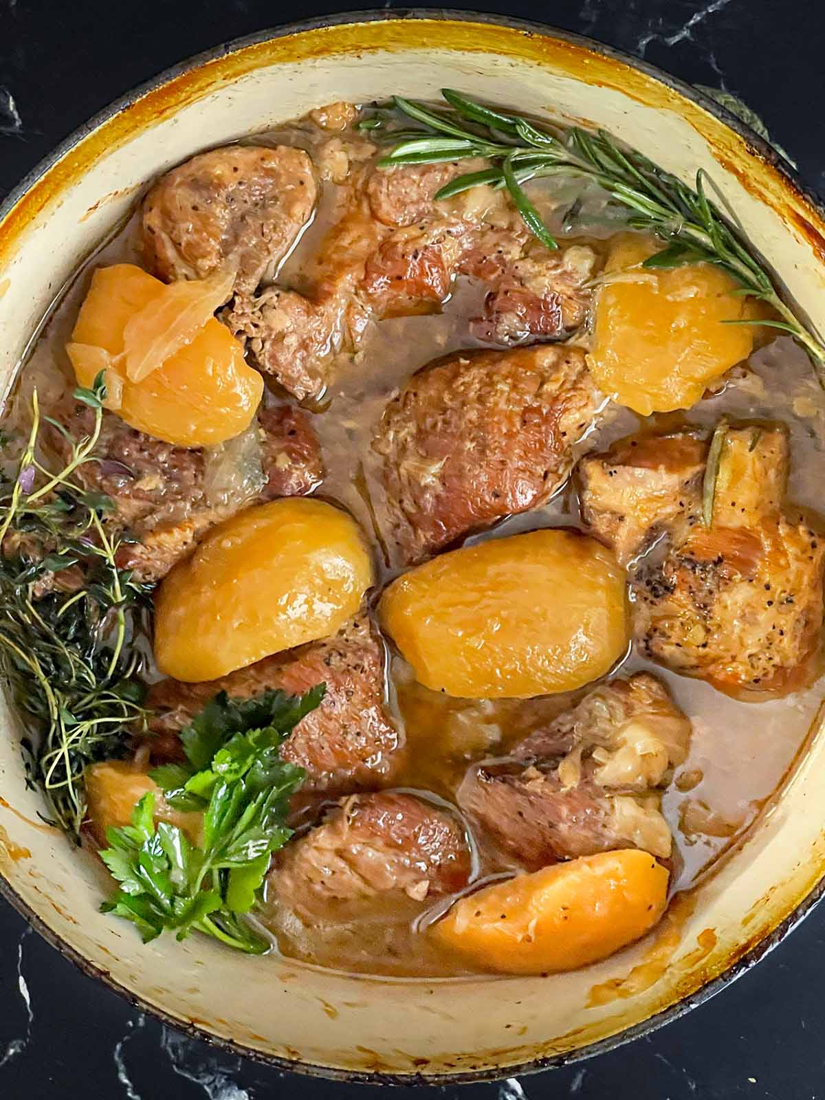 Apple cider braised pork shoulder in a dutch oven garnished with fresh rosemary and thyme.