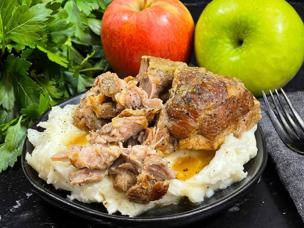 A plate of apple cider braised pork shoulder on a bed of mashed potatoes on a dark plate. Parsley and apples are in the background.