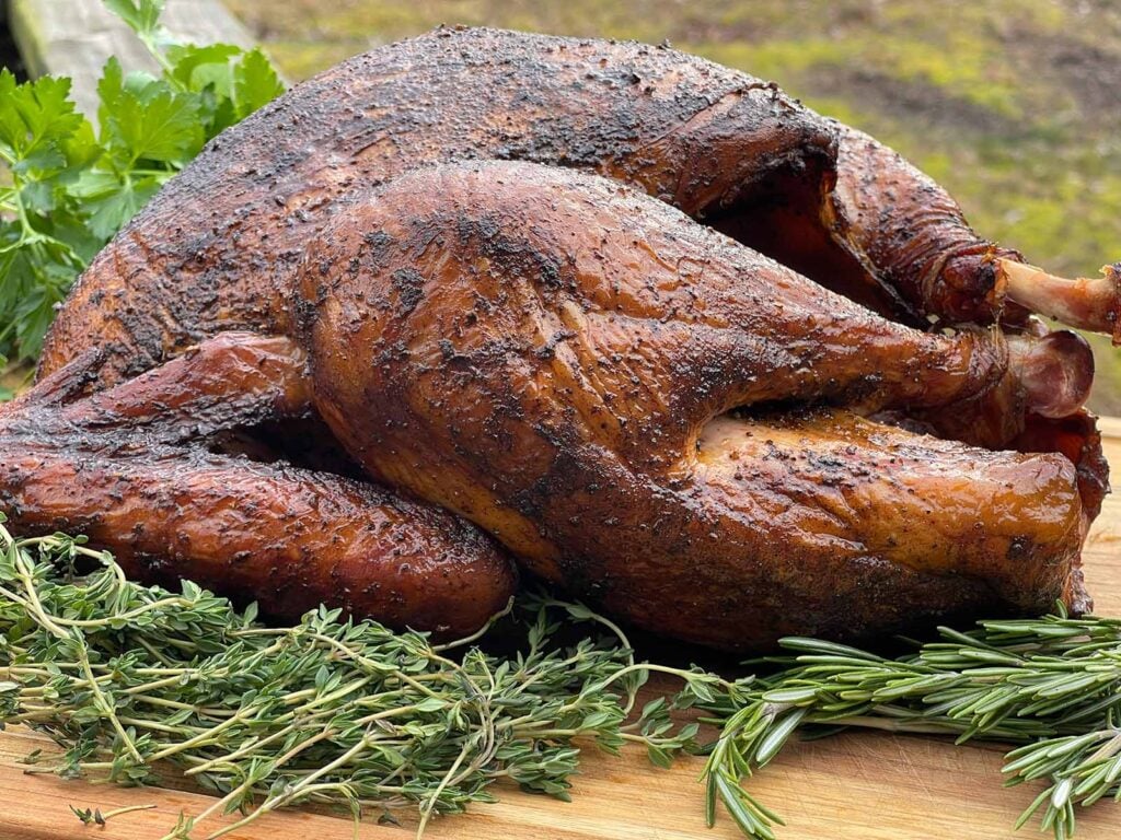 A whole smoked turkey on a wooden cutting board with fresh herbs.