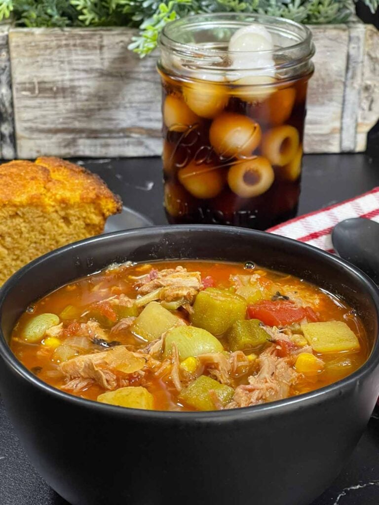 Brunswick Stew in a dark bowl with cornbread and a soda in the background.
