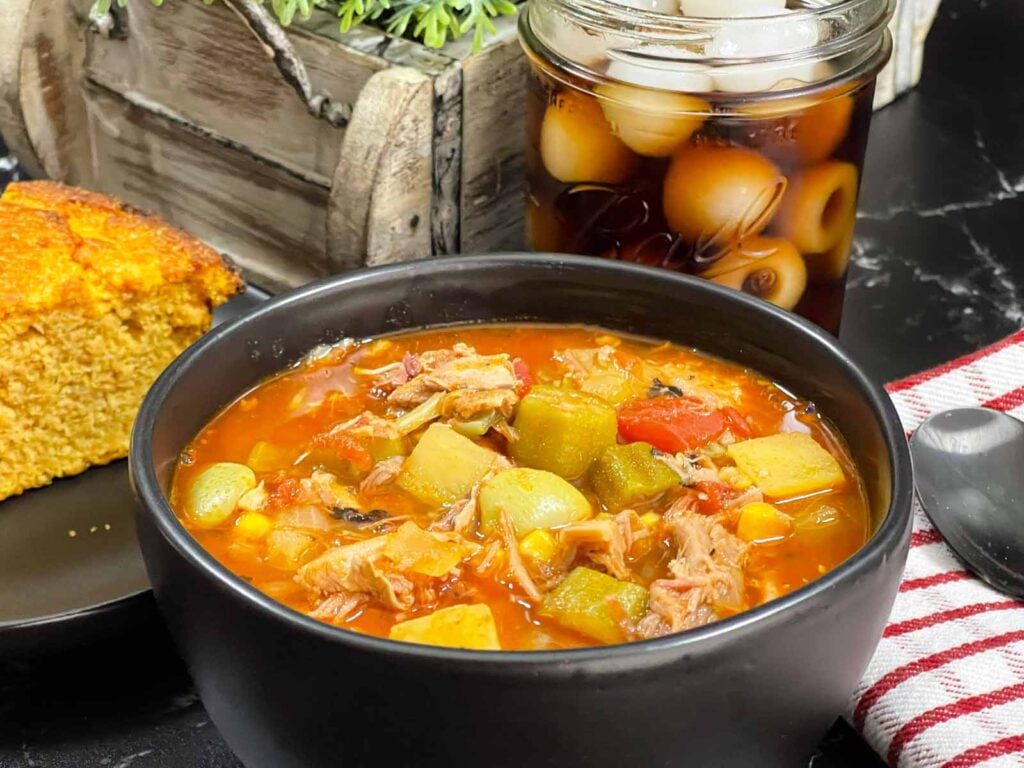 Brunswick Stew in a dark bowl with cornbread and a soda in the background.