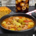 Brunswick Stew in a dark bowl with cornbread and a soda in the background.