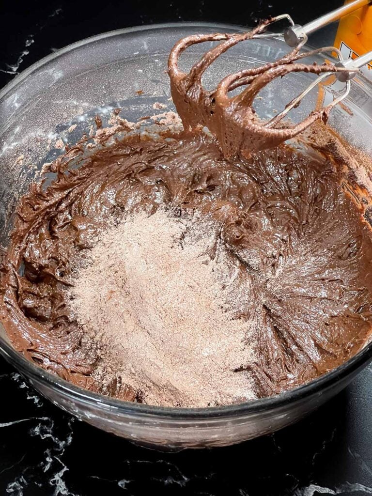 Dry ingredients being added to the wet ingredients in a glass bowl on a dark surface.