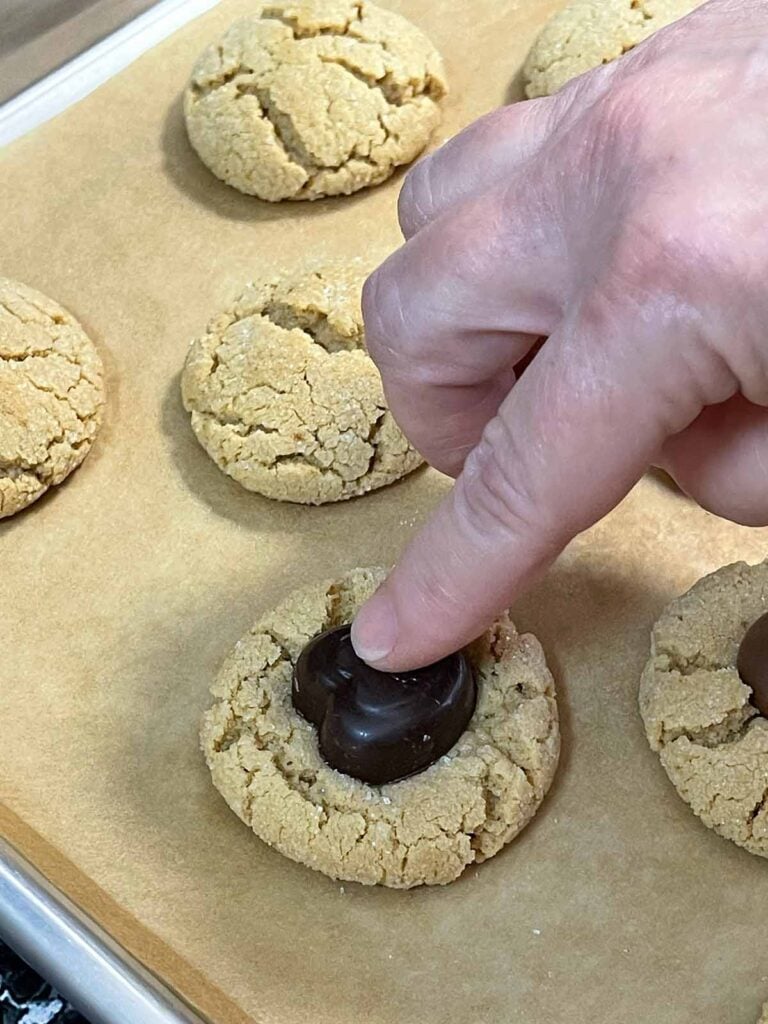 A chocolate heart being pressed into a hot peanut butter cookie on a baking sheet.