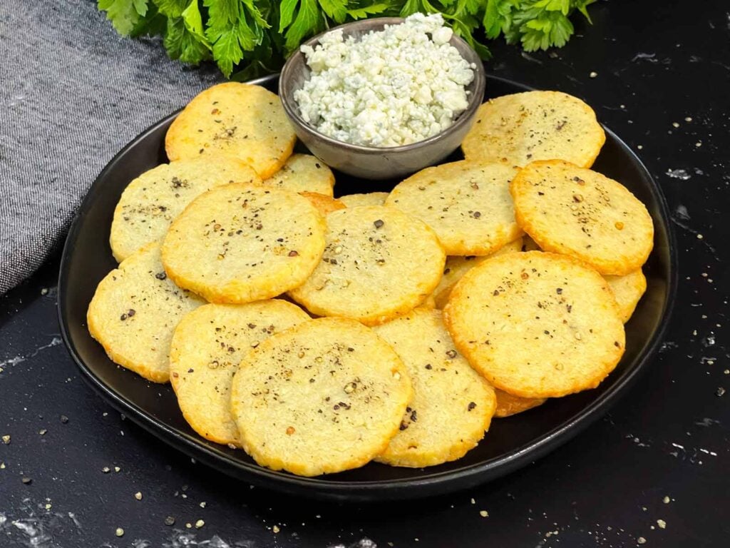 Blue cheese wafers stacked on a dark plate with a ramekin of crumbled blue cheese.