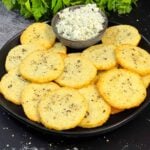 Blue cheese wafers stacked on a dark plate with a ramekin of crumbled blue cheese.
