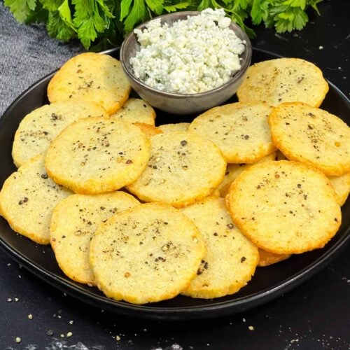 Blue cheese wafers stacked on a dark plate with a ramekin of crumbled blue cheese.