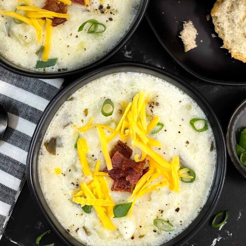 Creamy potato soup topped with diced bacon, shredded cheddar cheese, and diced green onions in a dark bowl.