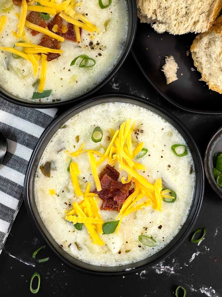 Creamy potato soup topped with diced bacon, shredded cheddar cheese, and diced green onions in a dark bowl.