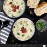 Creamy potato soup topped with diced bacon, shredded cheddar cheese, and diced green onions in dark bowls.