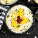 Creamy potato soup topped with diced bacon, shredded cheddar cheese, and diced green onions in dark bowls.