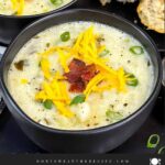 Creamy potato soup topped with diced bacon, shredded cheddar cheese, and diced green onions in dark bowls.