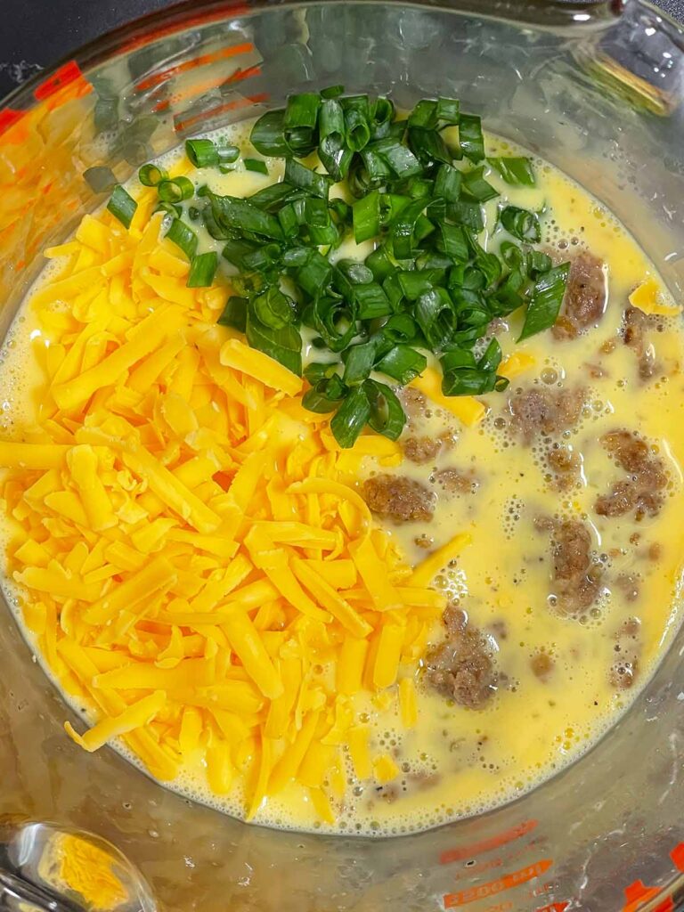 Shredded cheddar cheese, green onion, and sausage added to an egg scramble in a glass bowl.