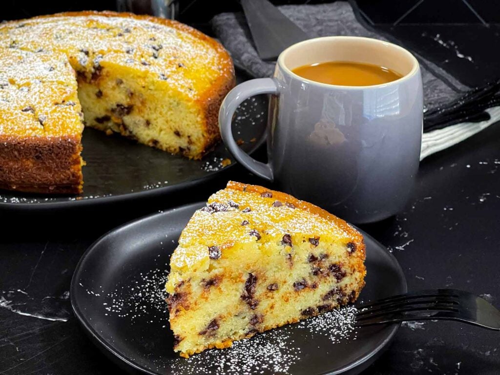 A slice of chocolate chip ricotta cake garnished with powdered sugar on a dark plate.