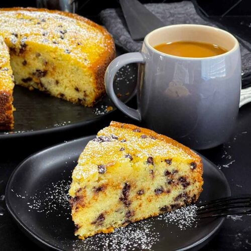 A slice of chocolate chip ricotta cake garnished with powdered sugar on a dark plate.