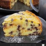 A slice of chocolate chip ricotta cake garnished with powdered sugar on a dark plate.