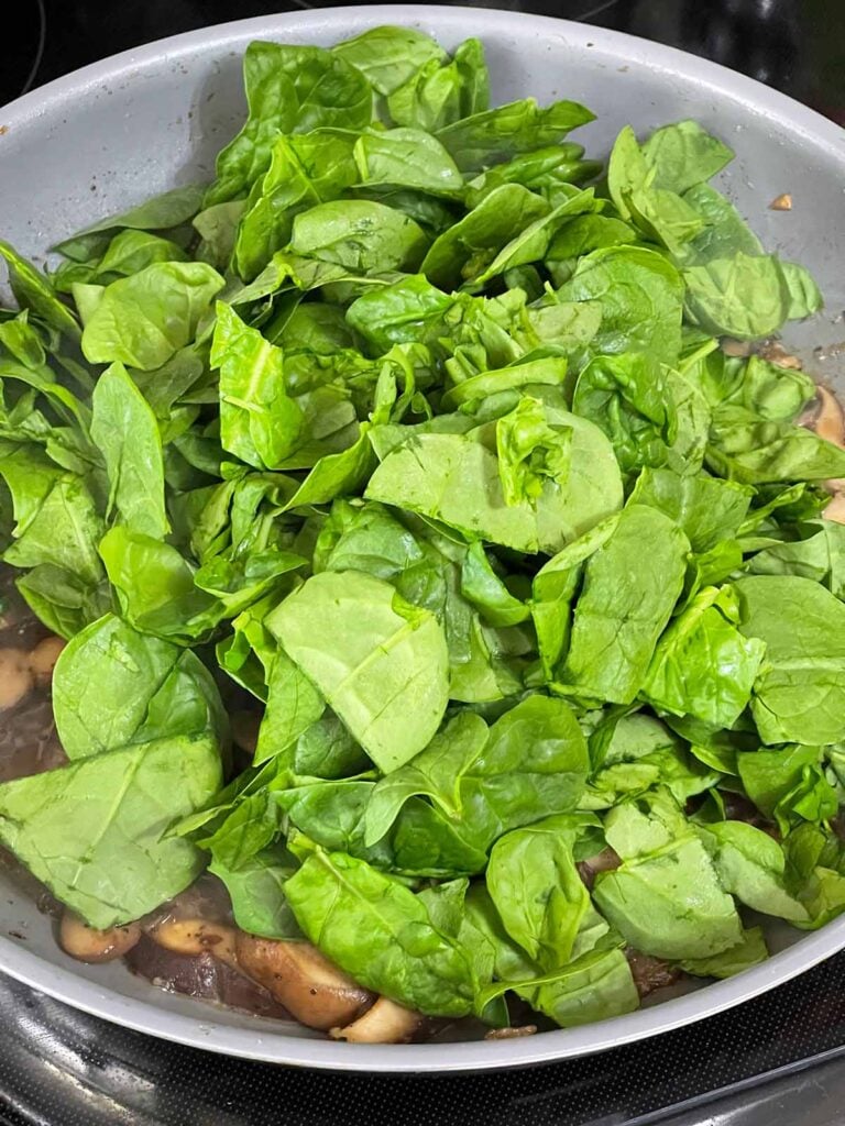 Baby spinach being added to a skillet with mushrooms.