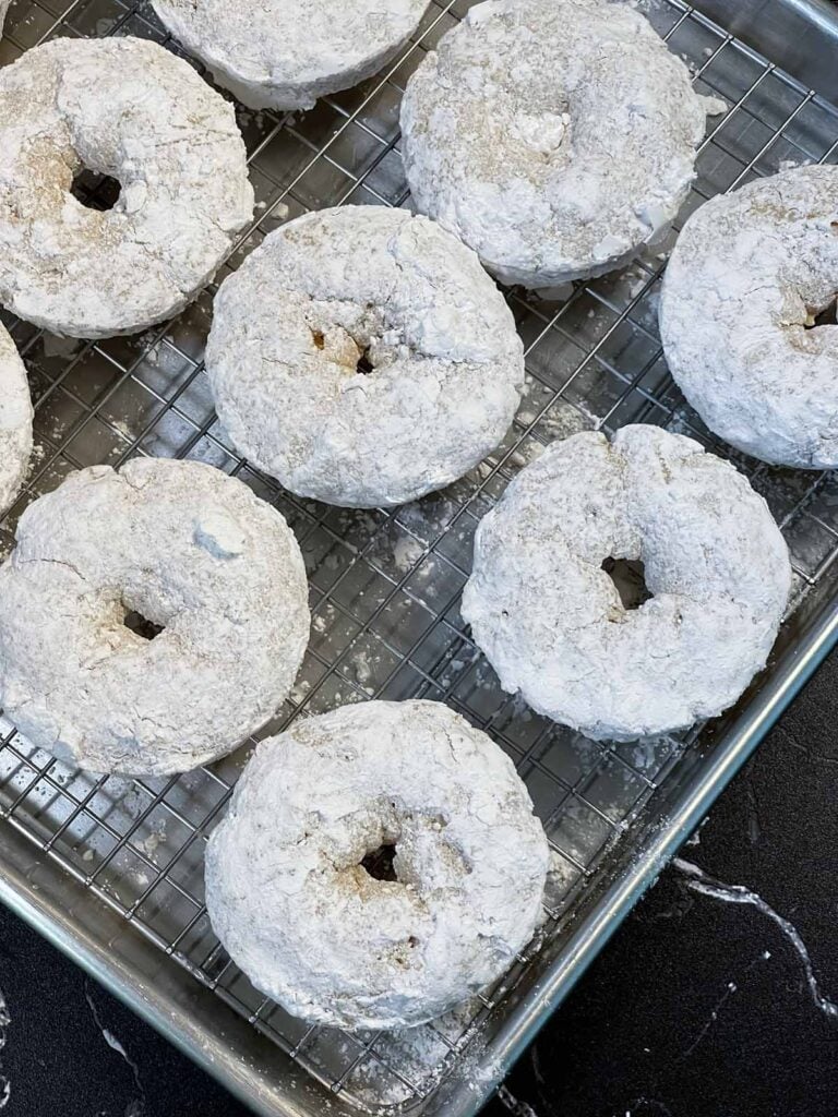Baked powdered sugar donuts on a wire rack over a baking sheet.