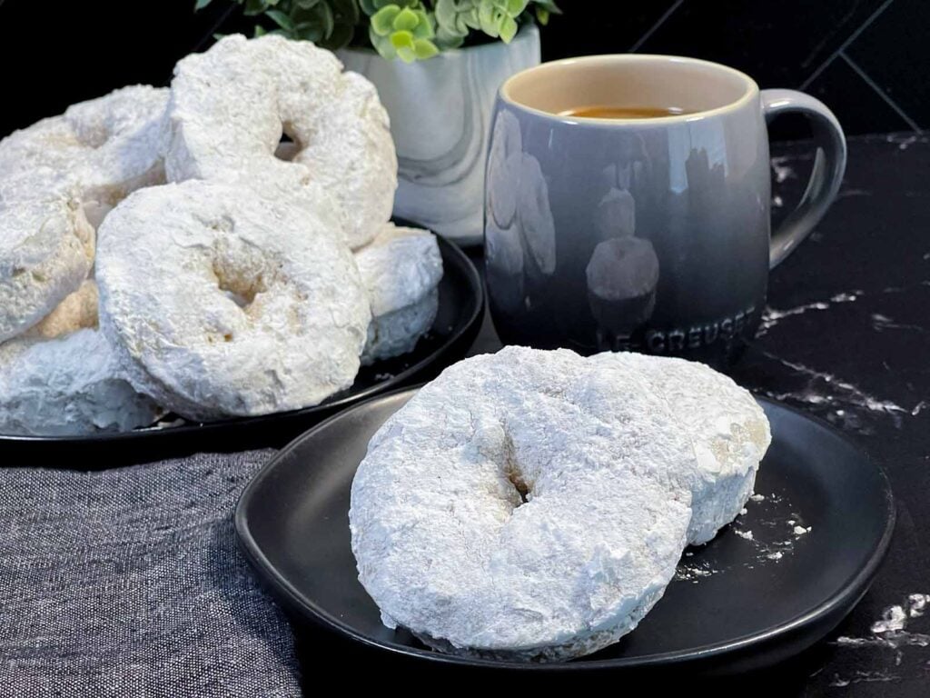 Powdered sugar coated baked donuts stacked on a plate with a soft, cake-like texture.