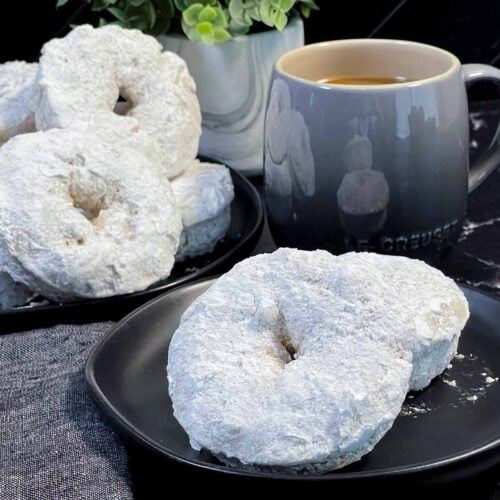 Powdered sugar coated baked donuts stacked on a plate with a soft, cake-like texture.