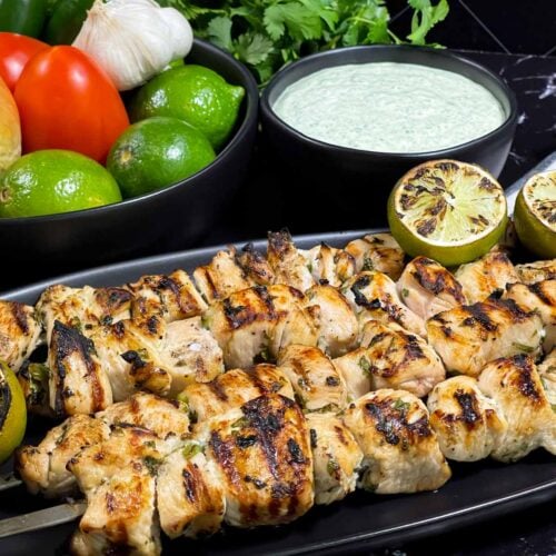Cilantro lime grilled chicken skewers on a dark plate with vegetables and dip in the background.