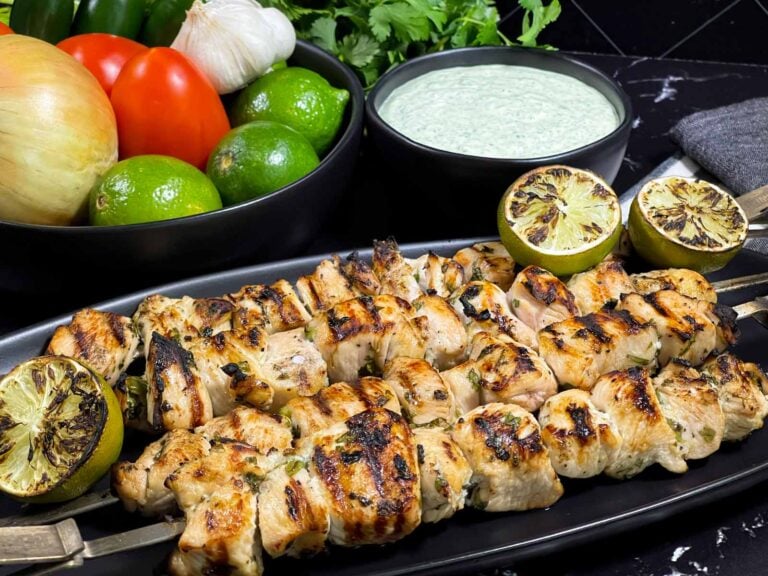 Cilantro lime grilled chicken skewers on a dark plate with vegetables and dip in the background.