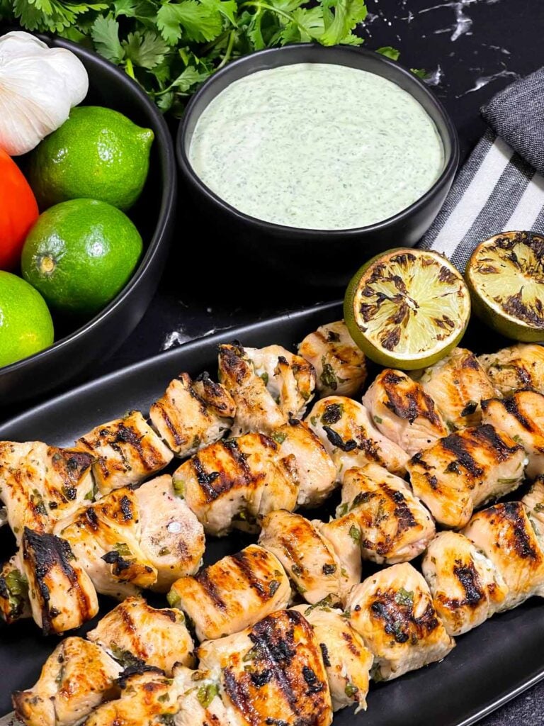 Cilantro lime grilled chicken skewers on a dark plate with vegetables and dip in the background.