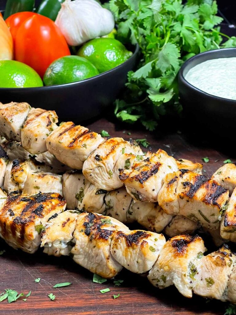 Cilantro lime grilled chicken skewers on a wooden cutting board with vegetables and dip in the background.