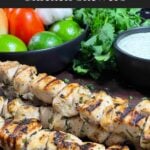 Cilantro lime grilled chicken skewers on a wooden cutting board with vegetables and dip in the background.