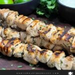 Cilantro lime grilled chicken skewers on a wooden cutting board with vegetables and dip in the background.