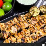 Cilantro lime grilled chicken skewers on a dark plate with vegetables and dip in the background.