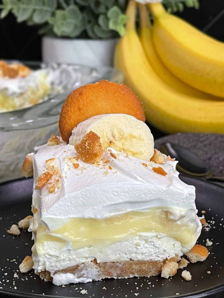 A slice of banana pudding lush on a dark plate garnished with crushed vanilla wafers.