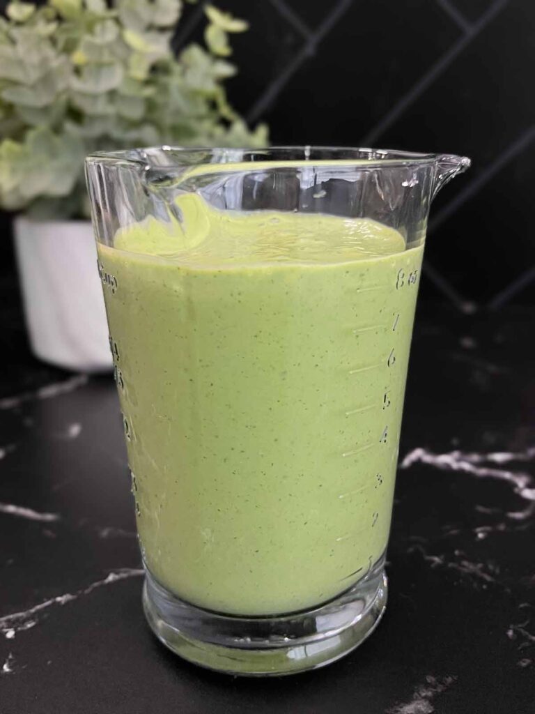 Green Goddess Salad dressing in a glass measuring cup on a dark surface.