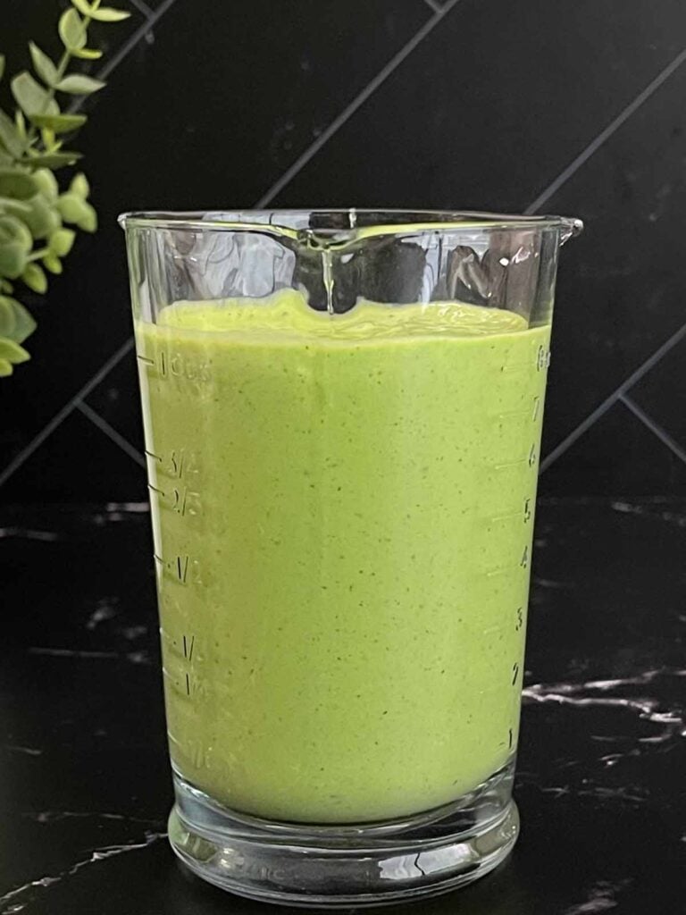 Green Goddess Salad dressing in a glass measuring cup on a dark surface.