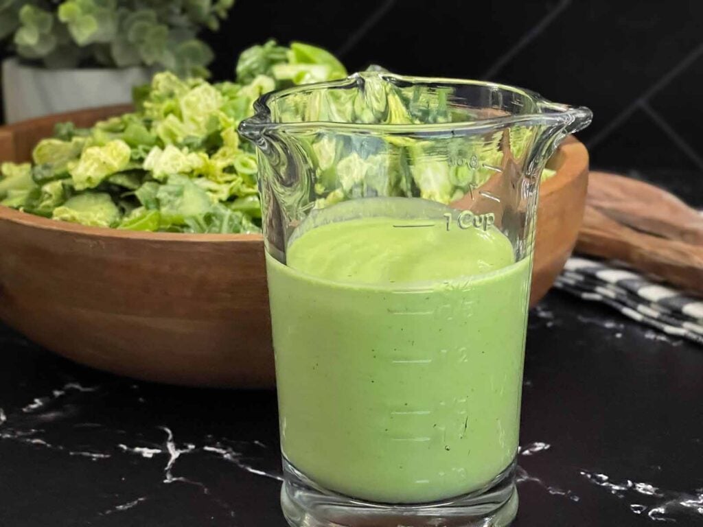 Homemade Green Goddess dressing in a glass measuring cup served in front of a fresh green salad.