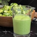 Homemade Green Goddess dressing in a glass measuring cup served in front of a fresh green salad.