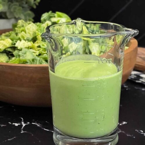 Homemade Green Goddess dressing in a glass measuring cup served in front of a fresh green salad.