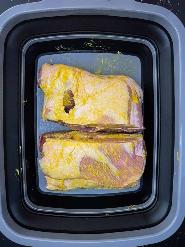 Two boneless pork butt halves coated with yellow mustard in a seasoning tub.