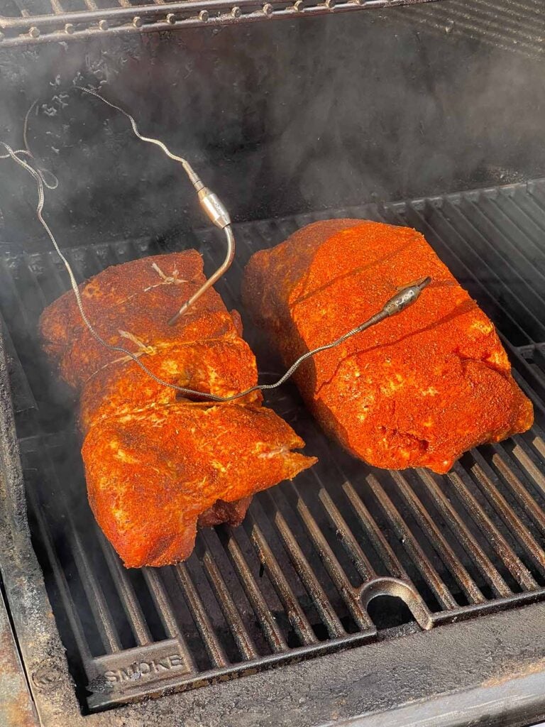Two seasoned boneless pork butt halves smoking at 250 degrees with temperature probes inserted.