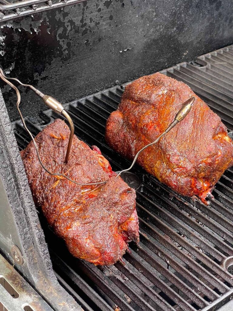 Boneless pork butt halves continuing to smoke as the bark darkens.