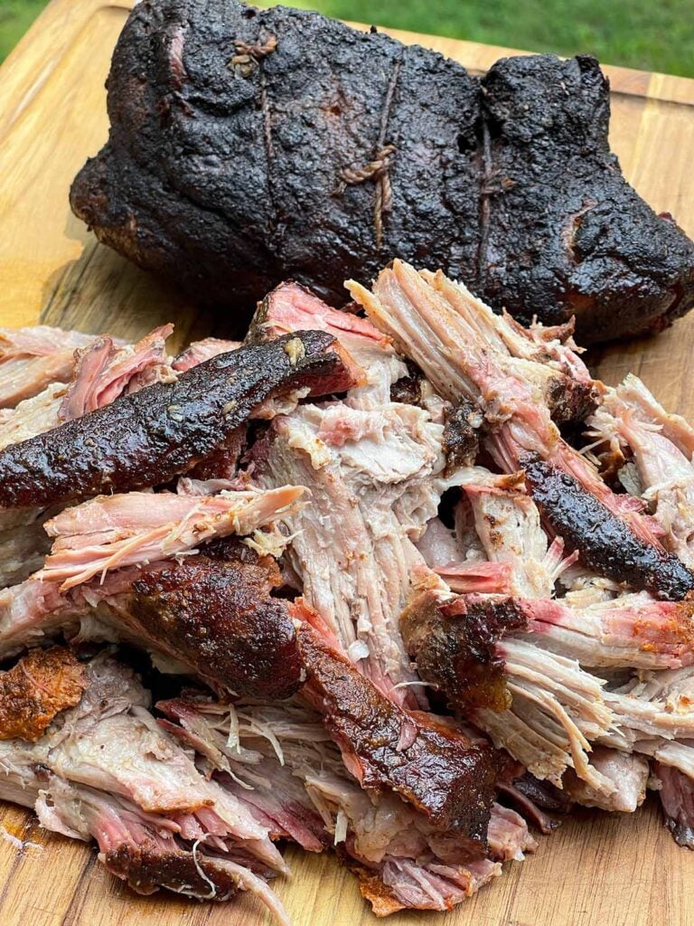 One smoked pork butt pulled apart beside the remaining whole half on a wooden cutting board.